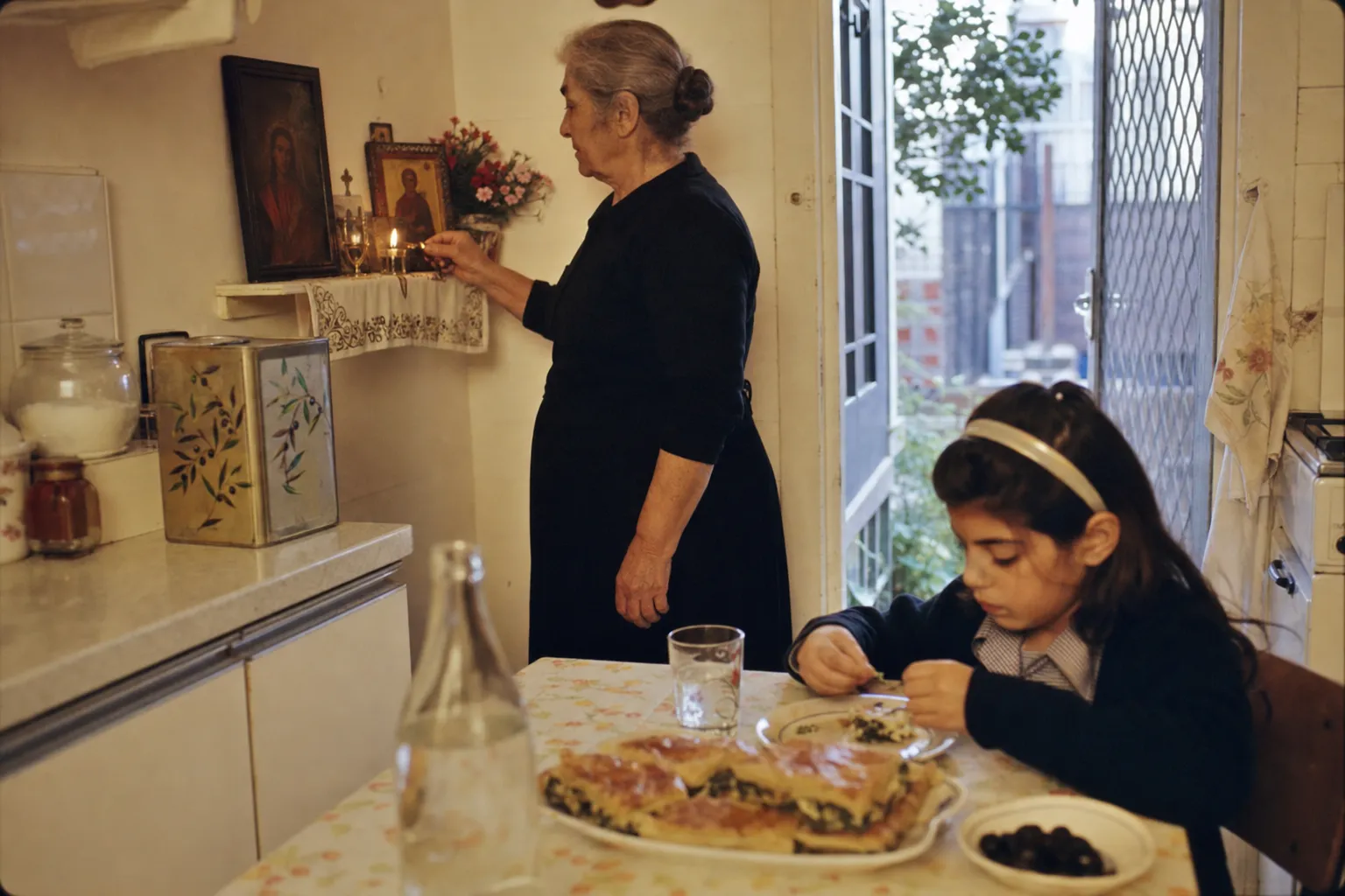 Greek-Australian yiayia lighting a candle in front of a Greek Orthodox icon corner in a Newtown terrace house
